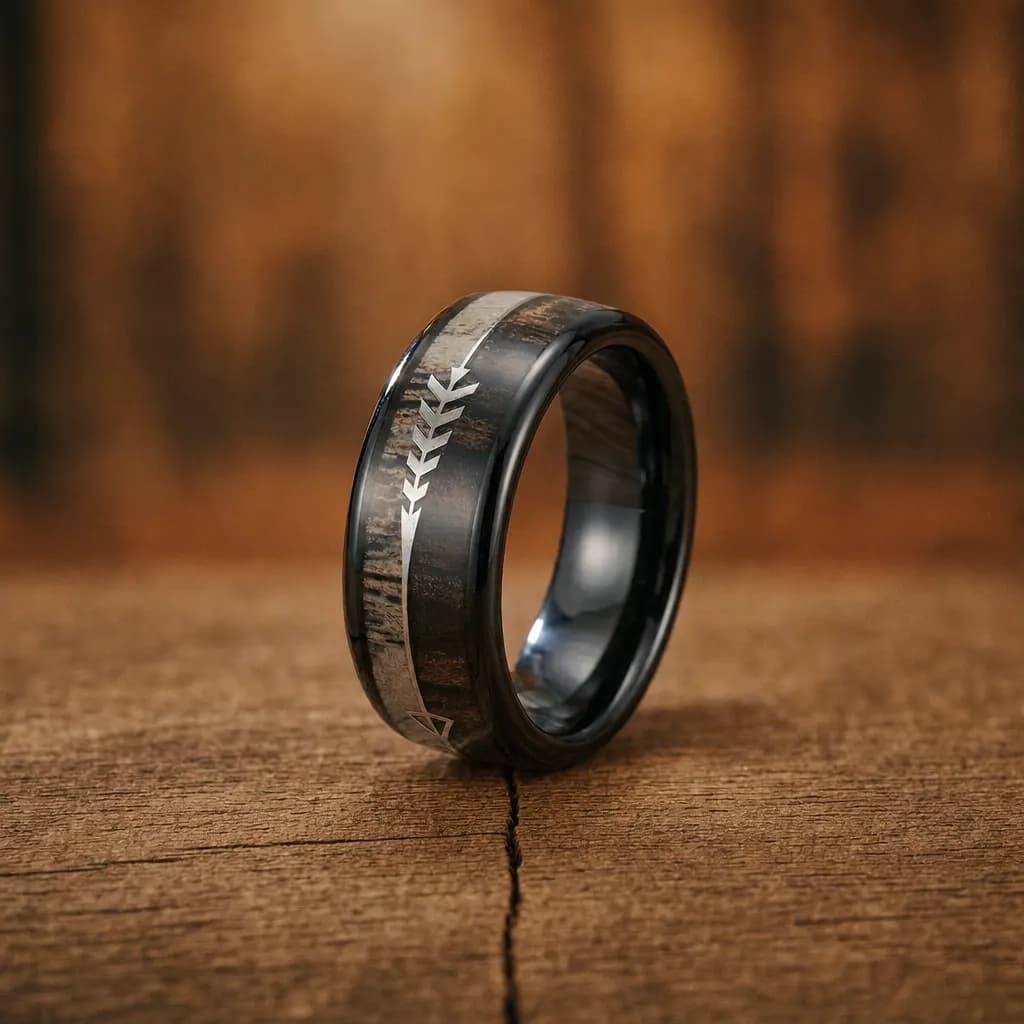 The Arrow, an 8mm polished black tungsten carbide band with an antler inlay, standing upright on a weathered oak plank