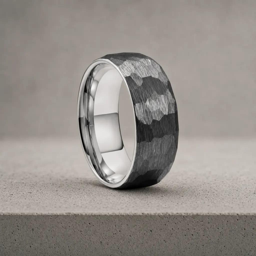 The Cobble, an 8mm hammered tungsten carbide band in gunmetal and silver, standing upright on a poured concrete ledge