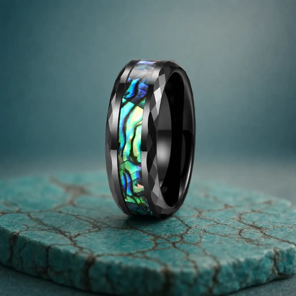 The Nautilus, an 8mm faceted tungsten carbide band in black, blue and green with an abalone shell inlay, standing upright on a turquoise veined mineral slab