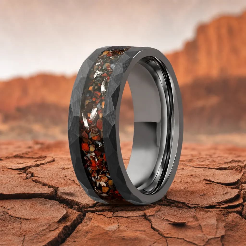 The Strata, an 8mm faceted tungsten carbide band in black and gunmetal with a dinosaur bone inlay, standing upright on cracked red desert earth