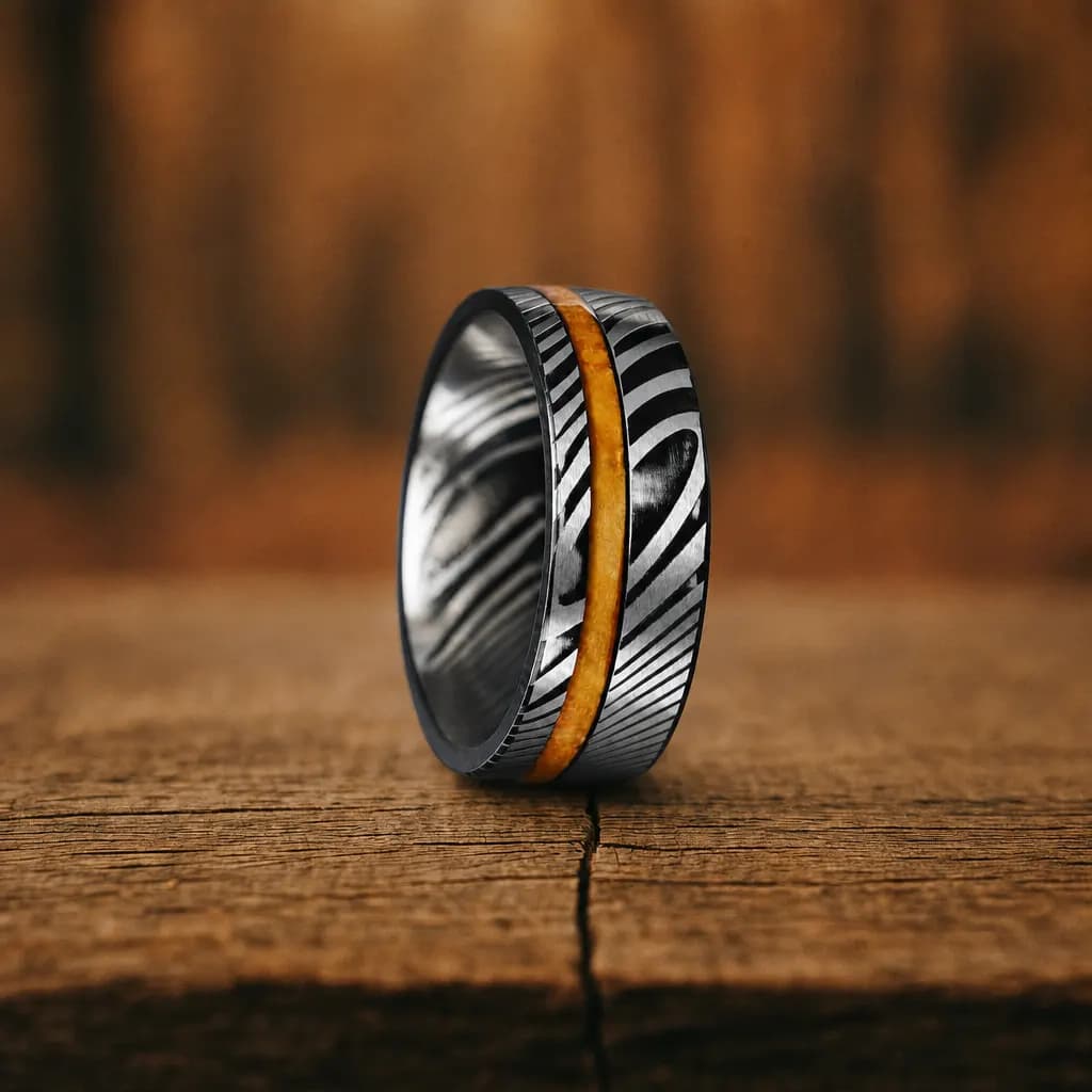 The Temper, an 8mm brushed damascus steel band in silver, black and gold with a wood inlay, standing upright on a weathered oak plank